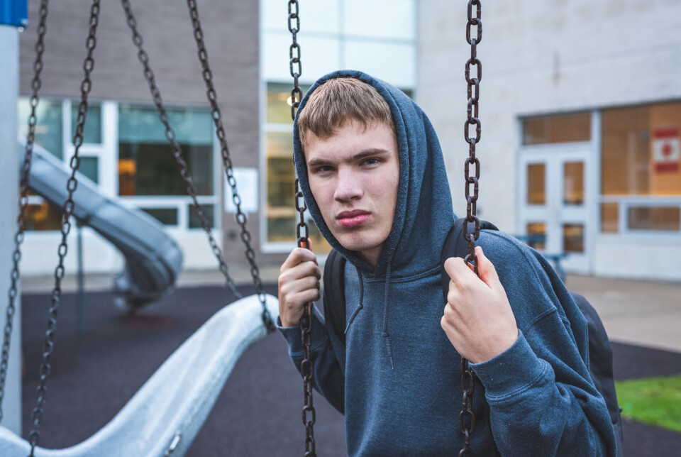 The image displays a sad teenage boy sitting on a swing outside of a school. He is reminiscing about when he was younger.