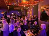 Publikum sitter ved småbord inne på en bar.